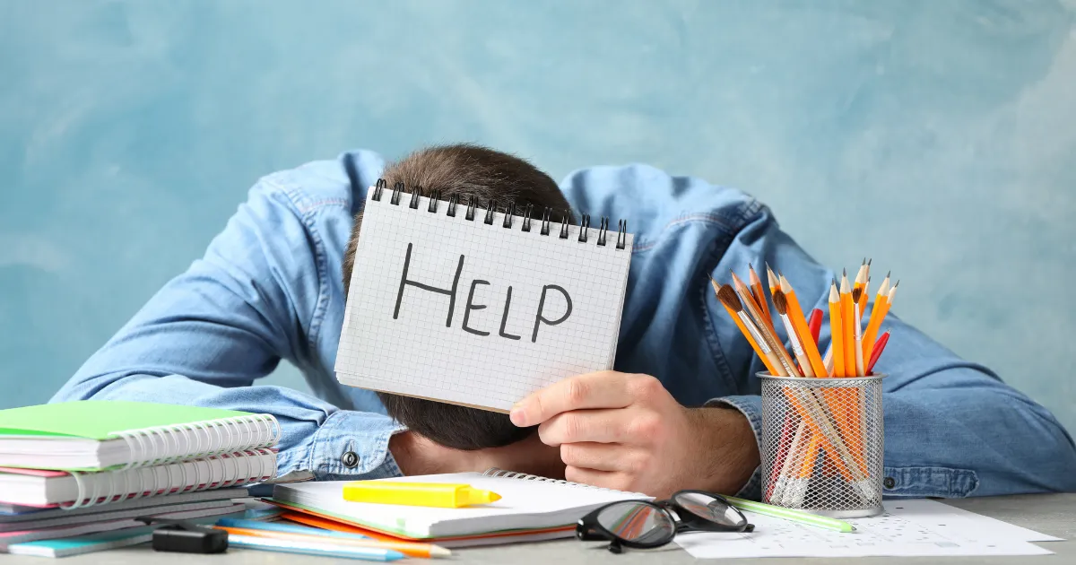 机に伏せて「HELP」と書かれたノートを掲げる疲れた男性